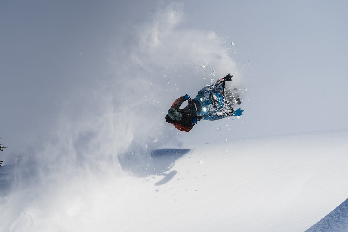 A snowmobile jumps a large snowbank with a rider airborne, kicking up a spray of snow in a bright, snowy outdoor setting.