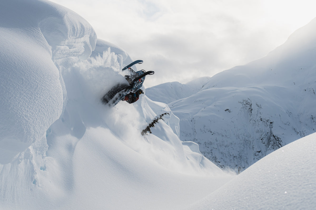 A snowmobile jumps high into the air while carving through deep powder snow on a steep mountain slope, with snow spraying around it and snowy peaks in the background.
