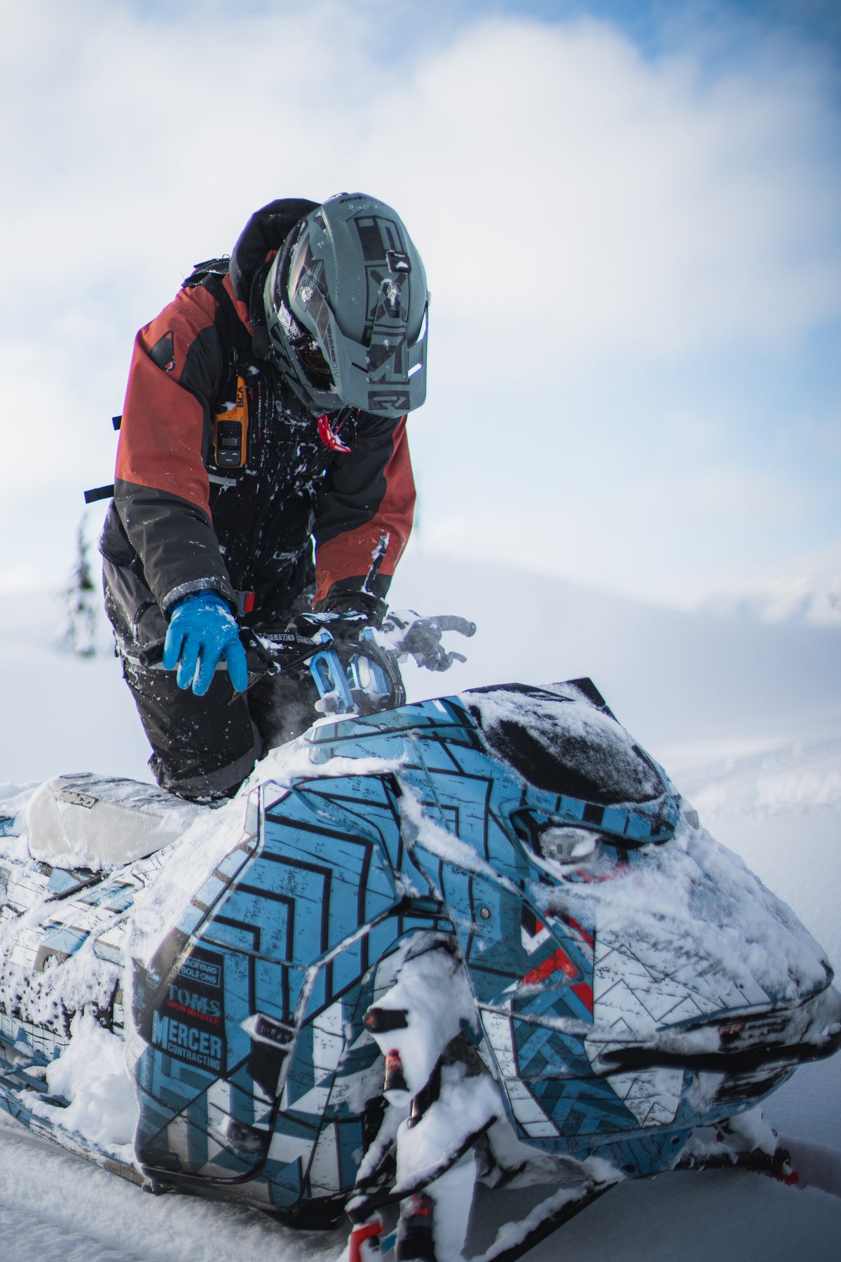 A snowmobile rider in winter gear, including a gray helmet, red and black jacket, and blue gloves, riding a snowmobile covered in snow in a snowy outdoor environment.
